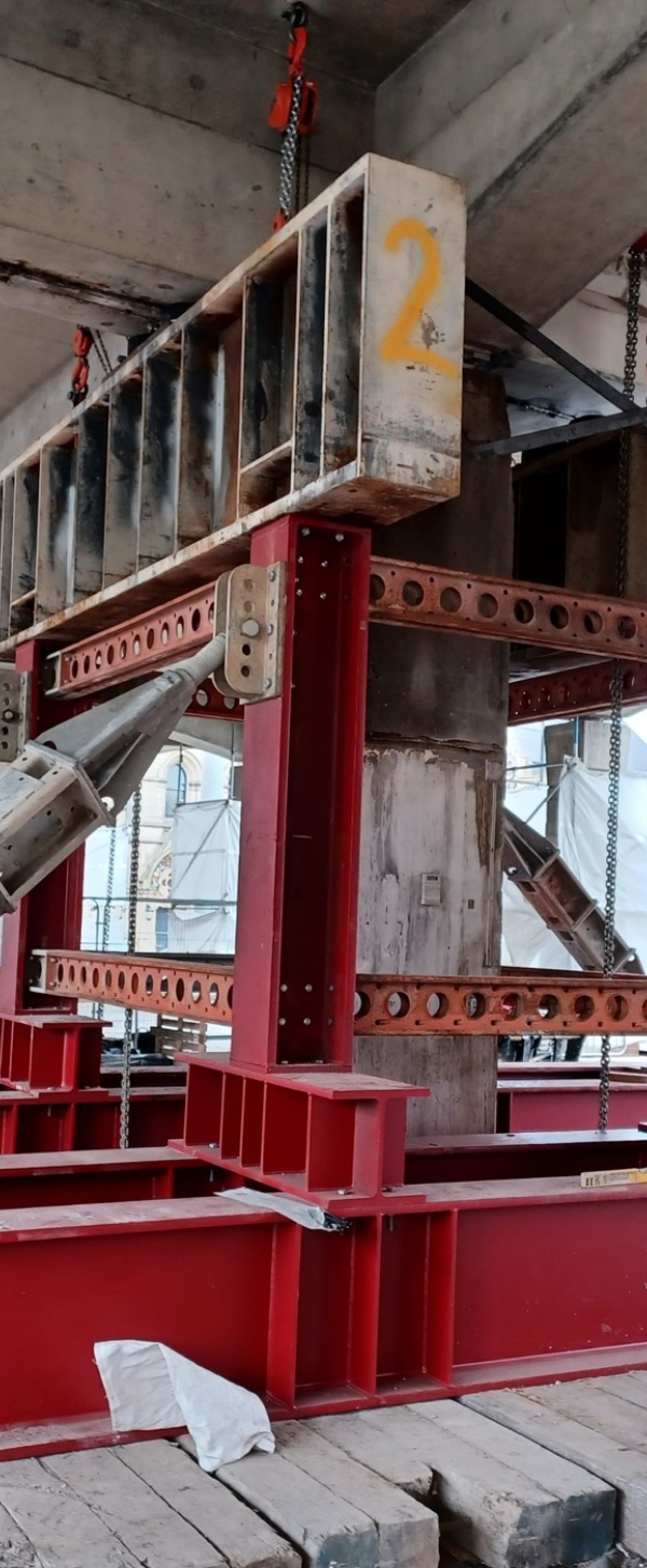 Structural steel support system at a construction site, with red steel beams forming a reinforced framework. The setup includes angled support struts bolted into place for stability, with chains hanging from the top beam to help distribute weight.