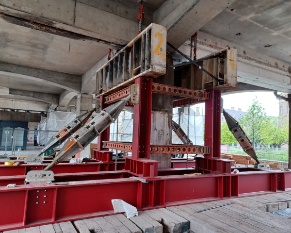 Structural steel support system at a construction site, with red steel beams forming a reinforced framework. The setup includes angled support struts bolted into place for stability, with chains hanging from the top beam to help distribute weight.