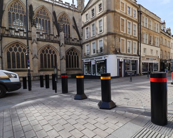 Black bollards with red and yellow bands on a cobbled street, in front of a historic building with Gothic windows and nearby shops