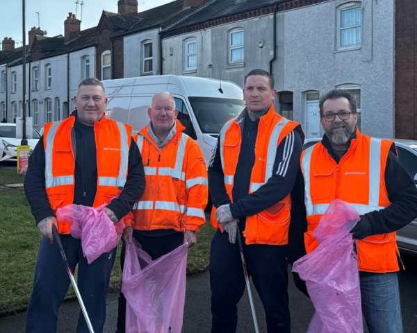 four men wearing orange hi-vis vests smiling at the camera holding purple bin bags litter picking