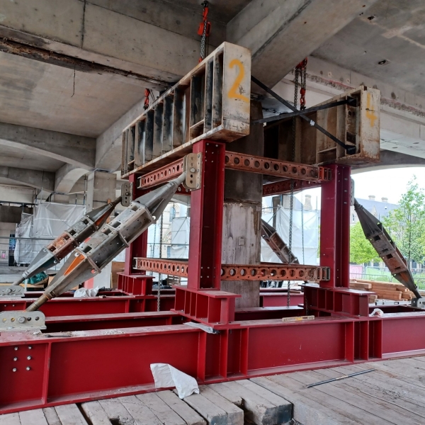 Structural steel support system at a construction site, with red steel beams forming a reinforced framework. The setup includes angled support struts bolted into place for stability, with chains hanging from the top beam to help distribute weight.