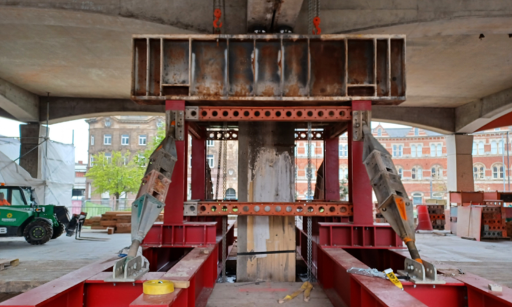 A front view of a temporary structural support setup under a concrete structure, featuring red steel beams arranged in a rectangular frame with bolted connections. Diagonal metal struts reinforce the structure, connecting the red framework to the upper support beam. 