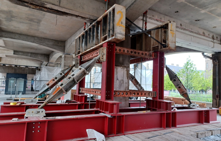 Structural steel support system at a construction site, with red steel beams forming a reinforced framework. The setup includes angled support struts bolted into place for stability, with chains hanging from the top beam to help distribute weight.